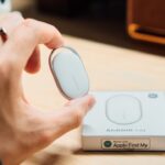 Rastreador inteligente ao lado de smartphone com tela de localização, fundo neutro e iluminação suave.