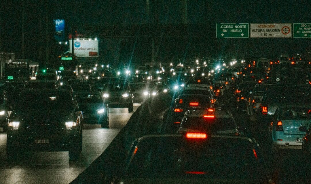 Semáforo suspenso sobre avenida movimentada ao anoitecer, com rastros de luz dos veículos ao fundo